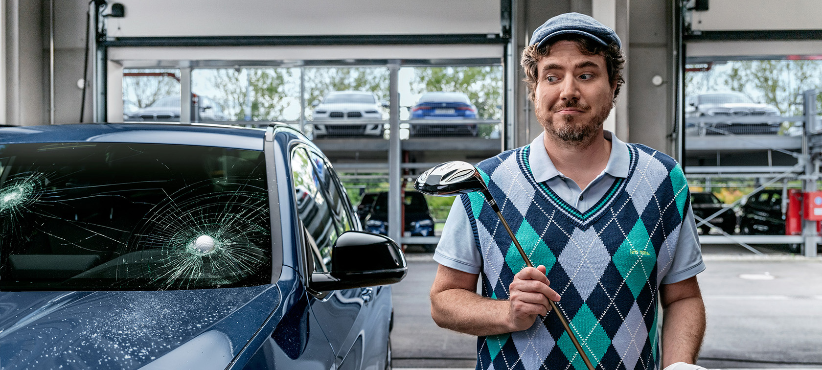 En man med golfklubba bredvid en BMW med en golfboll i vindrutan.