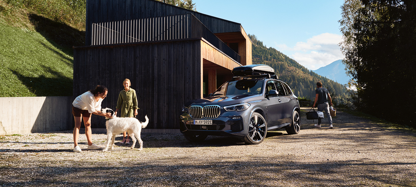 Familj med hund framför en BMW som parkerat framför ett hus i sommarmiljö.